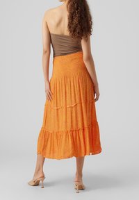 Žena s kudrnatými vlasy, oblečená v hnědém topu bez ramínek, oranžové midi sukni s volány a béžových vysokých podpatcích, stojící proti jednobarevnému pozadí.