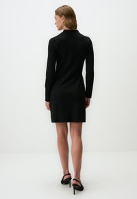 Robe noire à manches longues avec une silhouette ajustée, texture lisse et détail de couture à l'arrière. Associée à des sandales à talons noires.
