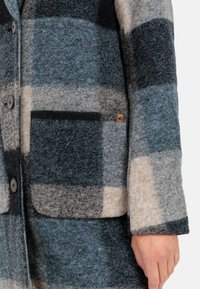 Grijze en blauwe geblokte wollen jas met een zak, zwarte accenten en knoopsluiting. Heeft een klein merklabel aan de zijkant.
