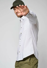 Wit shirt met een patroon van kleine kleurrijke geometrische ontwerpen, knoopsluiting aan de voorkant, lange mouwen, gecombineerd met groene broek en een pet.