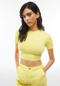 Keltainen lyhythihainen crop-top, joka on valmistettu pehmeästä materiaalista. Siinä on pyöreä kaula-aukko ja hillitty brändilogo edessä.