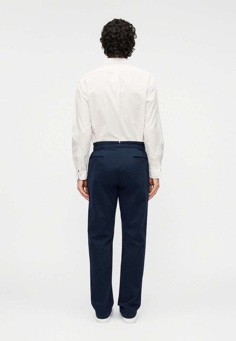 Pantalon marine en coton, avec une coupe droite, des poches latérales et des poches arrière boutonnées. Le modèle porte une chemise blanche à boutons.
