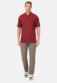Červené polo tričko s krátkým rukávem, zipovým límečkem a přední kapsou, v kombinaci s béžovými kalhotami a bílými teniskami s tmavými akcenty.