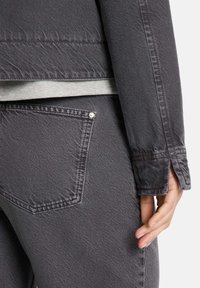 Graue Jeansjacke mit sichtbarer Textur, seitlichem Schlitz am Handgelenk und akzentuiertem Rückentasche. Weicher, leicht ausgebleichter Stoff.