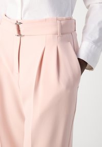 Pantaloni su misura rosa chiaro con pieghe e una cintura con fibbia in argento, realizzati in un tessuto morbido. Tasca laterale visibile.