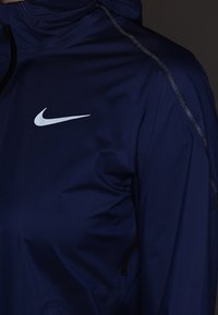 Veste de sport bleu foncé avec un logo Nike Swoosh réfléchissant sur la poitrine supérieure et une bordure réfléchissante le long de la couture de l'épaule.