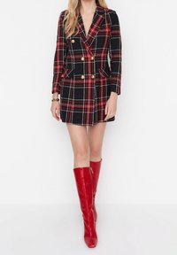 Femme portant une robe blazer à carreaux noir et rouge, croisée, avec des boutons dorés et des bottes rouges brillantes montantes jusqu'au genou.