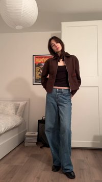 Bruine cropped jas met ritssluiting, zwarte tanktop en losse blauwe spijkerbroek. Zwarte platte schoenen, minimale accessoires, zachte binnenverlichting.