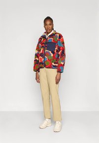 Färgstark fleecejacka med ett livfullt mönster av cirklar i rött, blått, grönt och rosa, med en enfärgad marinblå ficka och dragkedja.