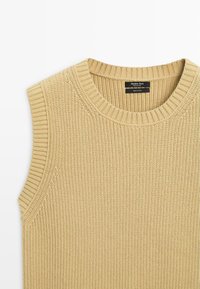 Pull sans manches en maille de coton beige avec encolure ronde côtelée et emmanchures, motif de points verticaux texturés, et étiquette de marque noire à l'intérieur du col.