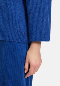 Sweter z głębokiego niebieskiego koloru w prążki, o luźnym kroju, z mankietami na rękawach i małym metalowym akcentem na końcu, zestawiony z spodniami z sztruksu.