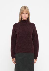 Bordowy sweter z dzianiny o wysokim dekolcie, w oversize'owym kroju i teksturowanym wzorze. W połączeniu z ciemnoszarym spódnicą z długimi rękawami.
