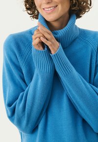 Modra turtleneck pulover iz mehke pletene tkanine. Ima rebraste manšete in ovratnik, z ohlapnim krojem in strukturiranimi vzorci pletenja.
