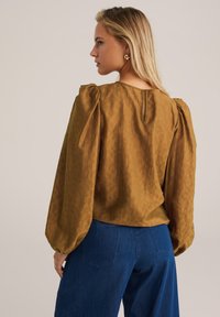 Bronzefarbene Bluse mit Puffärmeln, strukturiertem Stoff und einem Schlüssellochverschluss hinten. Kombiniert mit blauen weit geschnittenen Jeans.