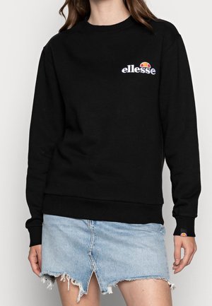 Frau trägt ein schwarzes ellesse-Sweatshirt mit Logo auf Brust und Bündchen, kombiniert mit einem hellblauen, ausgefransten Jeans-Minirock mit Frontschlitz.