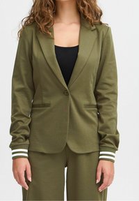 Blazer vert olive à un bouton, coupe ajustée, deux poches avant et poignets rayés. Le tissu semble lisse et extensible.