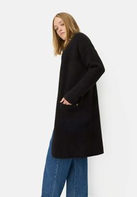 Schwarze, lange Strickjacke mit entspannter Passform, strukturiertem Stoff, seitlichen Taschen und gerippten Bündchen, getragen über locker sitzenden blauen Jeans.