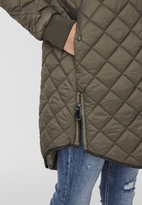 Tikkukuvioinen, oliivinvihreä takki, jossa on vetoketju edessä, ribbikantit ja hieman pidempi takapuoli. Pehmeä, sileä pinta, jossa on geometrinen kuvio.