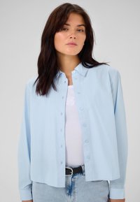 Camicia button-up azzurro chiaro realizzata in tessuto morbido, con maniche lunghe e vestibilità rilassata. Indossato sopra un top bianco a costine e jeans.