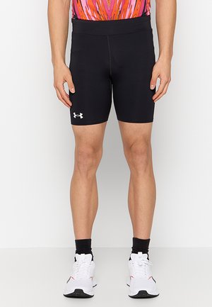 Uomo che indossa pantaloncini compressivi neri Under Armour fino al ginocchio, top rosa e arancione a fantasia, calzini neri e scarpe da ginnastica bianche, in piedi con le mani rilassate.