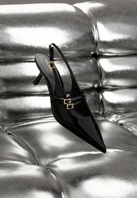 Scarpa slingback in vernice nera con punta affusolata, due accentuazioni in oro con fibbie e un tacco alto e sottile, esposta su una superficie argentata testurizzata.