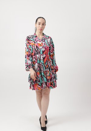 Mujer de pie vestida con un vestido largo de manga larga de capas con patrones en rosa, naranja, azul verdoso y negro, con tacones negros, sobre un fondo blanco liso.