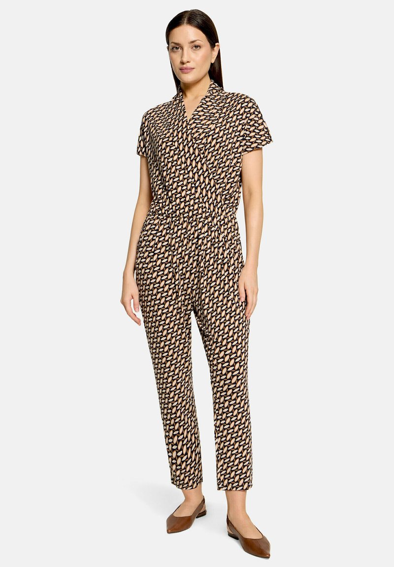 Femme debout portant une combinaison à manches courtes avec un motif de feuilles marron et beige et des chaussures plates marron sur un fond uni.