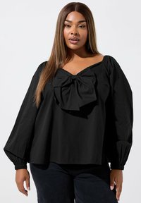 Svart blus med puffärmar och en stor rosett detalj vid halsringningen. Tillverkad av lättviktigt tyg, med en lös och avslappnad passform.