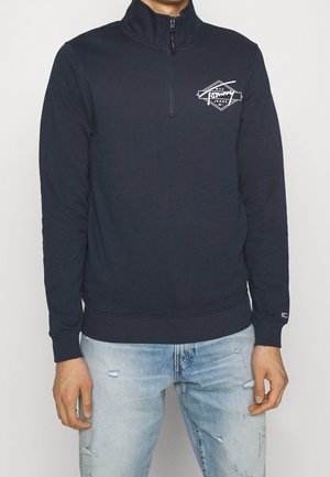 Mężczyzna w granatowej bluzie zapinanej na zamek do połowy wysokości z białym logo na piersi i jasnoniebieskich podniszczonych dżinsach, stoi z ramionami swobodnie opuszczonymi wzdłuż ciała.