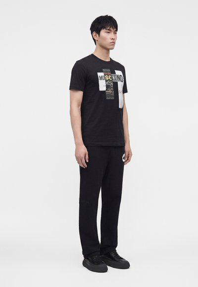 Νέος άνδρας που φοράει μαύρο graphic t-shirt Moschino, μαύρο παντελόνι με τσεπάκι με φερμουάρ και μαύρα αθλητικά παπούτσια μπροστά σε ένα απλό λευκό φόντο.