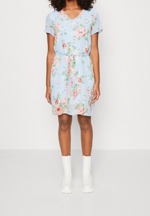 Femme portant une robe fleurie bleu clair à manches courtes et une ceinture à la taille, assortie à des bottes blanches épaisses, debout devant un fond uni.