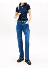 Mörkblå kortärmad topp med logotyp, högmidjade blåjeans och svarta läderstövlar. Modellen håller en vit väska och står framför en vit bakgrund.