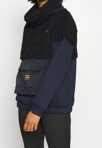Maglione navy con parte superiore in fleece nero e testurizzato, colletto a cowl e tasca frontale con etichetta. Polsini e bordo elasticizzati.