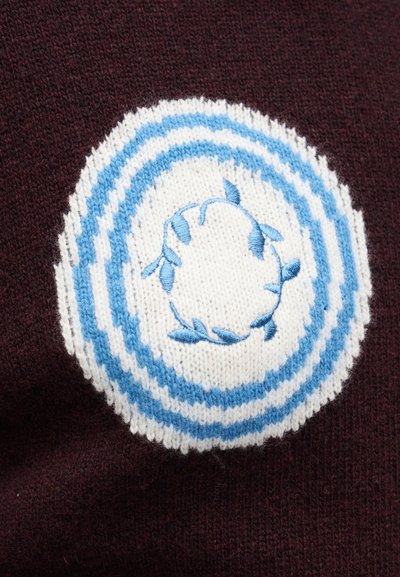 Strikket genser i mørk burgunder med en rund lapp med blå og hvite striper og blomsterbroderi i midten.
