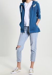 Blå zipjacka med huva, vit t-shirt under, ljusblå rivna jeans och guldskor med vita sulor.