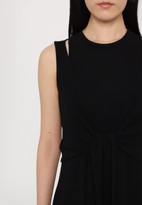 Femme portant une robe noire sans manches avec un détail noué à l'avant et une bretelle intérieure noire légèrement visible sur l'épaule gauche.