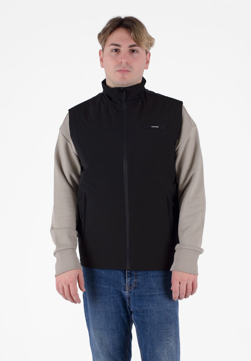 Gilet nero con colletto alto, zip frontale e tasche laterali. Le maniche sono grigio chiaro con polsini a coste. Presenta un piccolo dettaglio del logo sul petto.