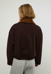Felpa corta marrone con colletto in pelliccia sintetica marrone, caratterizzata da una vestibilità rilassata e orlo a coste. Indossata con pantaloni felpa grigi.