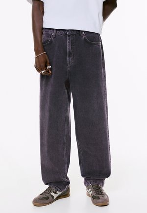 Jeans larghi color viola scuro in denim, dotati di tasche frontali e chiusura con bottone in vita, abbinati a sneakers grigie.
