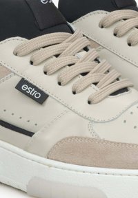 Beige leren sneakers met zwarte accenten, gestructureerde suède details, ronde veters en een witte rubberen zool. "Estro" logo op de zijkant.