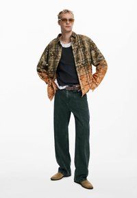 Chemise à carreaux dans des teintes de marron et d'orange, t-shirt noir à manches longues en dessous, jeans larges vert foncé, ceinture marron et chaussures marron clair.