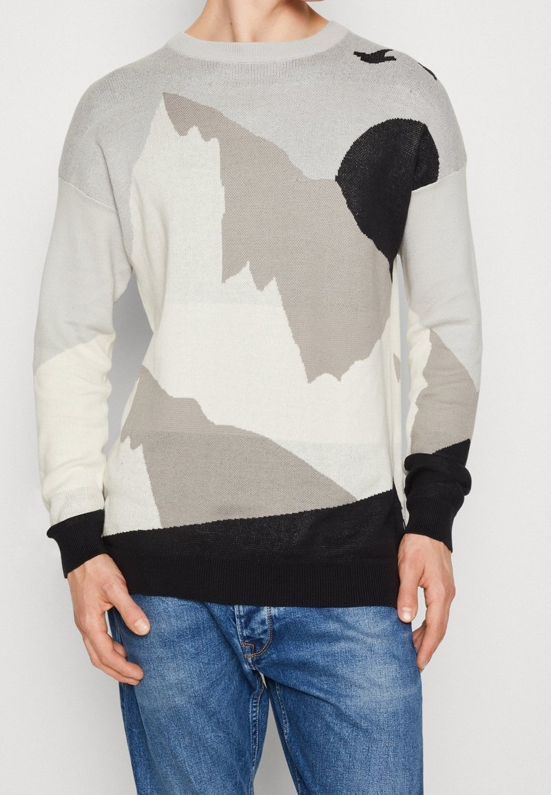 Homme portant un pull en tricot à motif abstrait gris, blanc, noir et beige avec un jean bleu, debout devant un fond uni.