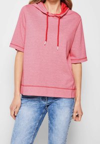 Kvinde iført en rød kortærmet hoodie med snore og lyseblå blegede jeans, stående foran en ensfarvet baggrund.