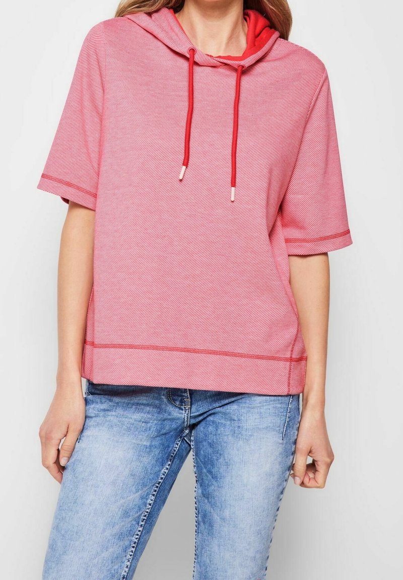 Kvinde iført en rød kortærmet hoodie med snore og lyseblå blegede jeans, stående foran en ensfarvet baggrund.