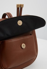 Ruskean tekstuurisen nahkabackpackin etutasku, musta kangasläppä ja kultaiset magneettiklipit, esitelty osittain auki valkoisen pinnan päällä.