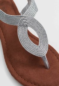 Correa de plata trenzada con acento circular sobre la suela de sandalia de ante marrón. Diseño plano con estilo minimalista, adecuado para uso casual.