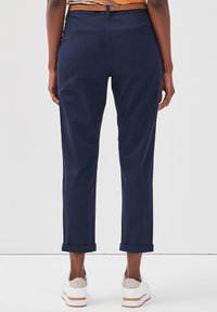 Pantalons bleu marine à rouleaux avec une texture lisse, deux poches arrière et une ceinture tressée marron, associés à des baskets blanches.