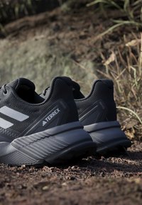 Pár čiernych bežeckých topánok Adidas Terrex na trail, ktoré odpočívajú na štrkovej ceste s trávou a kameňmi v pozadí.