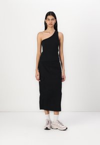 Black asymmetric one-shoulder top paired with a long black cargo skirt featuring two side pockets and light-colored athletic shoes.