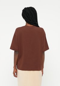 Camiseta castanha de manga curta em um leve tecido de algodão com um corte descontraído e gola redonda, vista de costas contra um fundo neutro.
